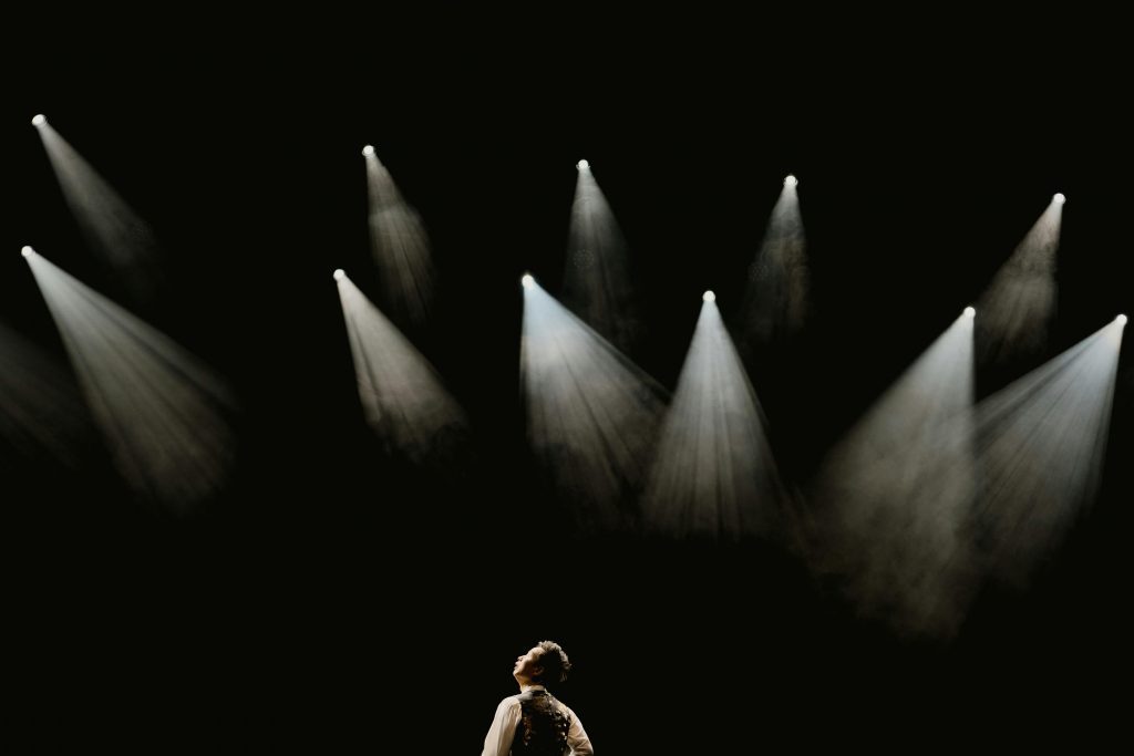 A solo performer stands in a spotlight on a dark stage during a dramatic moment.