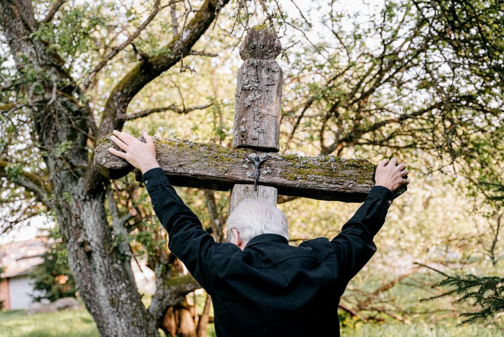 pexels-photo-8964629-8964629 An elderly man embraces a wooden cross, expressing grief and religion in an outdoor setting.