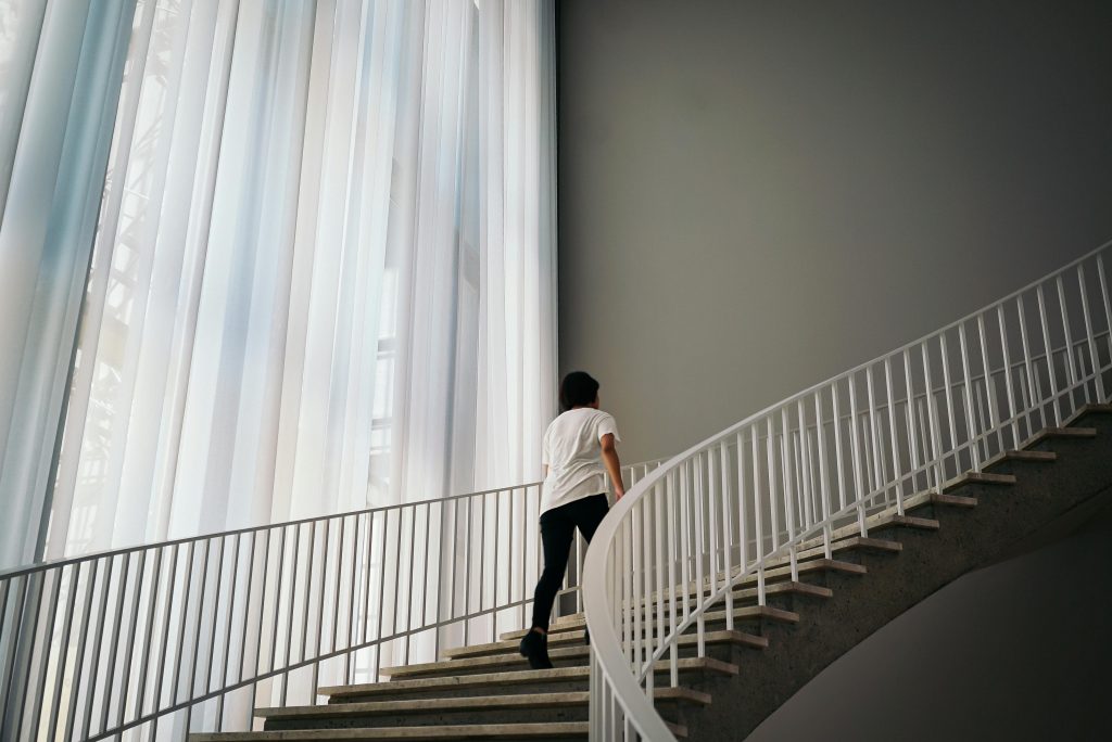 A person ascends a modern indoor staircase with soft window light filtering through curtains.