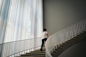 A person ascends a modern indoor staircase with soft window light filtering through curtains.