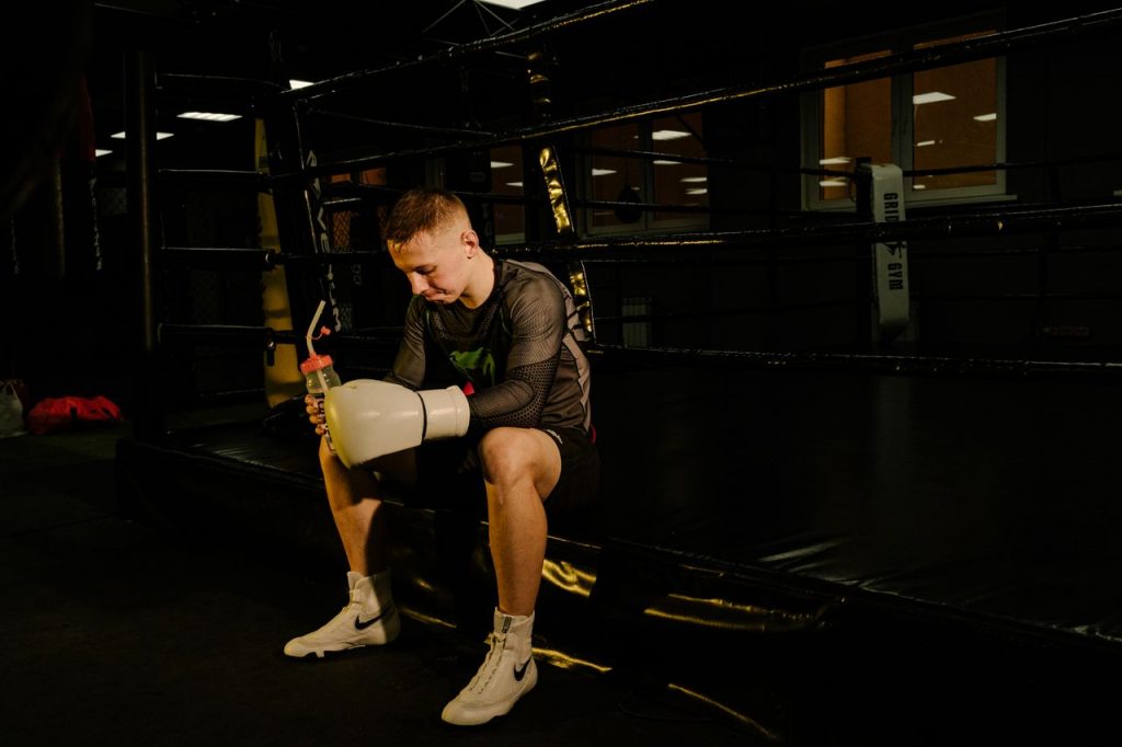 Young male boxer sitting in the ring holding gloves and a water bottle, taking a break.